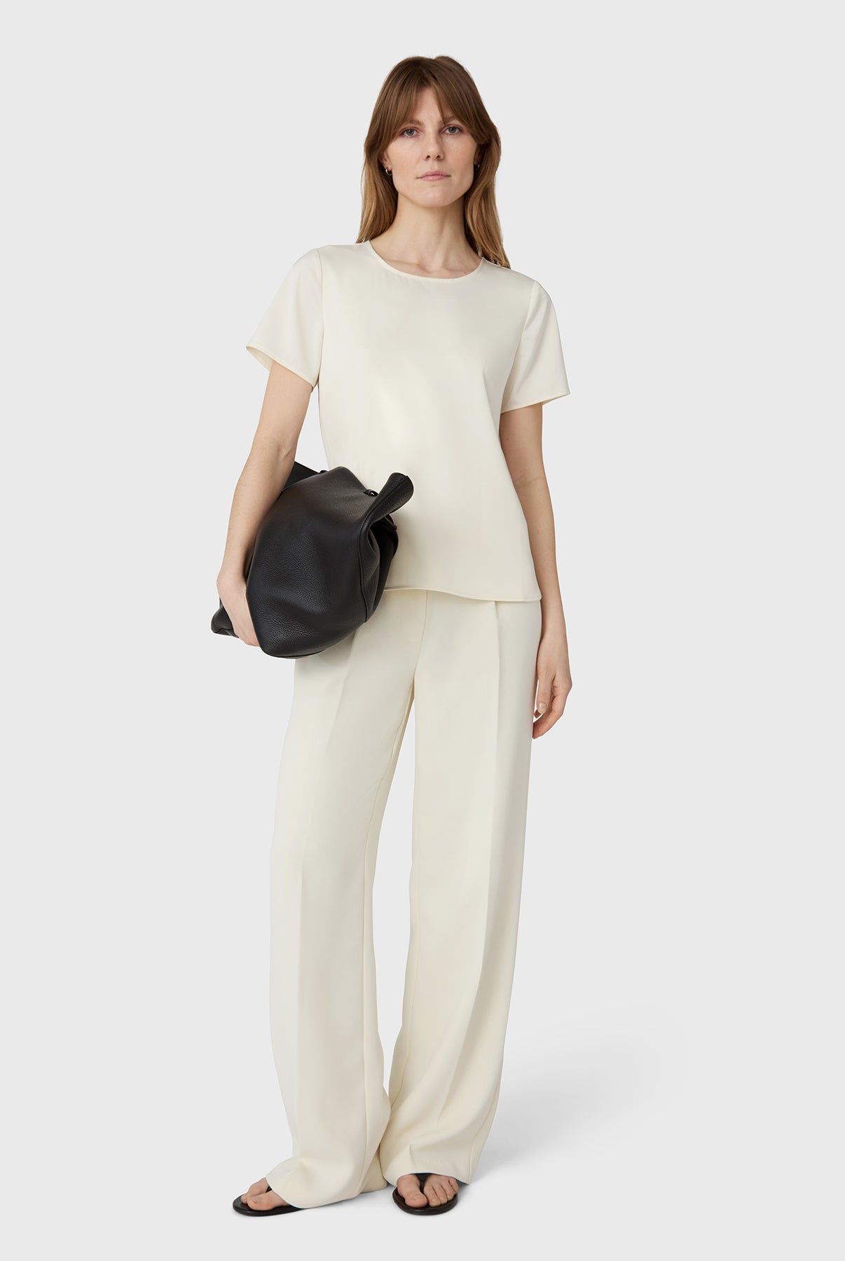 Woman wearing a white top and beige pants holding a black bag on a white background
