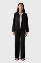 A woman standing, wearing black trousers and a black jacket with a V-neckline.