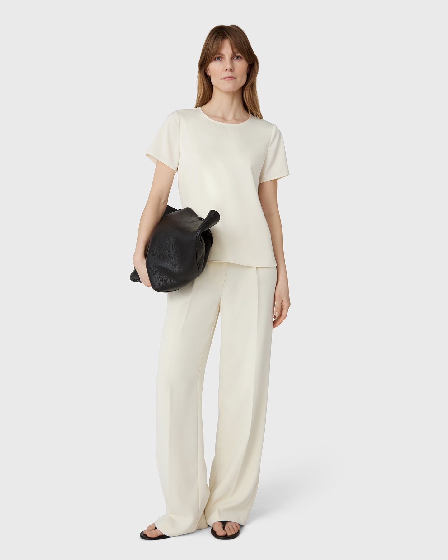 Woman wearing a white top and beige pants holding a black bag on a white background